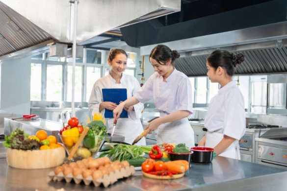 Trois jeunes cuisinières en tenue blanche s'exercent ensemble dans une cuisine professionnelle pour apprendre à cuisiner sous la supervision d'un chef.