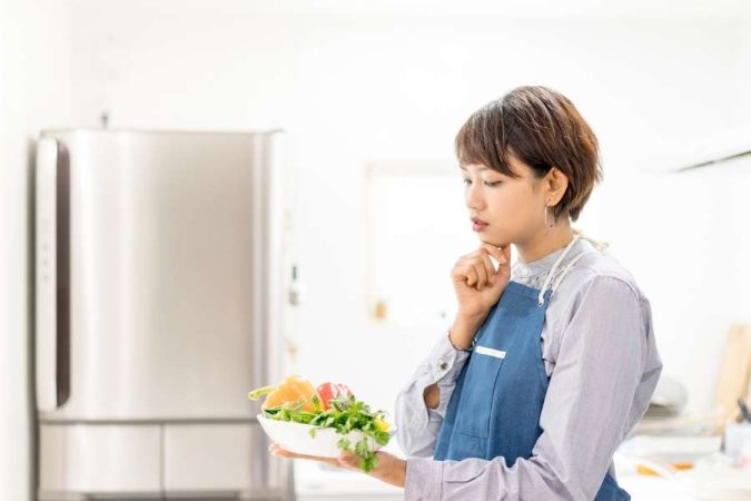 Une jeune femme en tablier bleu tient un bol de légumes frais et réfléchit à sa prochaine recette pour apprendre à cuisiner.