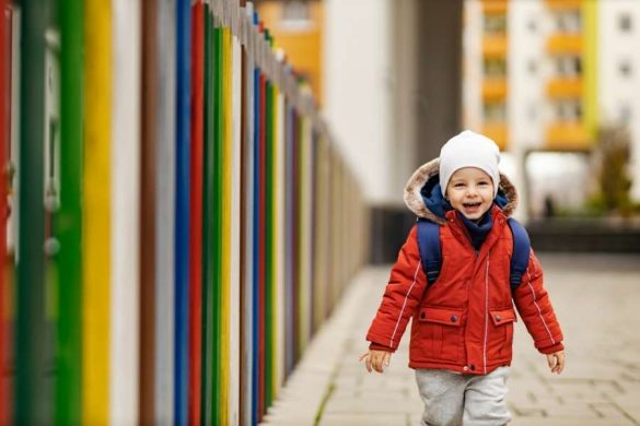 Enfant souriant portant une doudoune rouge et un sac à dos bleu marine, marchant près d'une clôture colorée.