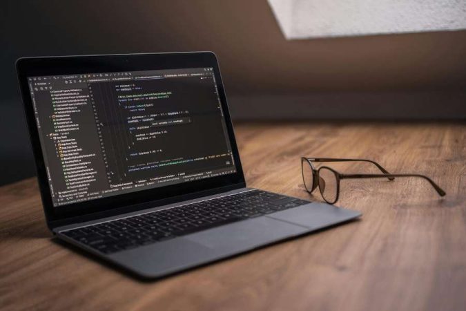 Un ordinateur portable ouvert affichant du code informatique complexe sur son écran, posé sur un plancher en bois, avec une paire de lunettes à côté.