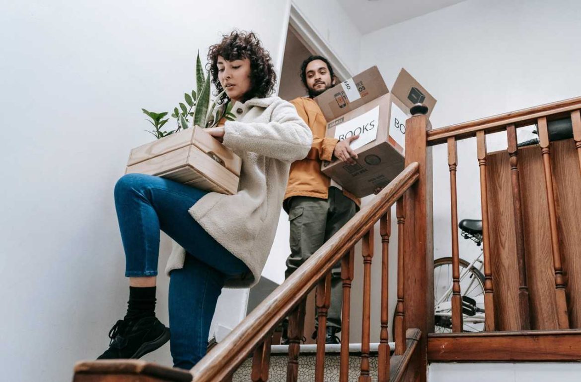Un couple descend un escalier en bois lors d'un déménagement. La femme au premier plan porte une cagette en bois contenant des plantes vertes, tandis que l'homme à l'arrière-plan transporte des cartons de livres