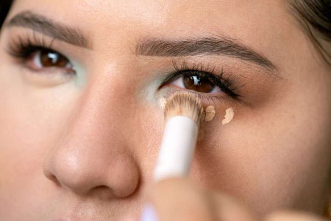 Gros plan sur une femme qui applique délicatement du maquillage sous ses yeux à l'aide d'un pinceau et d'un correcteur anti-cernes.