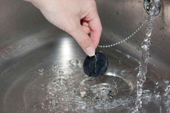 Une main tenant un bouchon en caoutchouc noir relié à une chaînette au-dessus d'un évier en inox où l'eau s'écoule.
